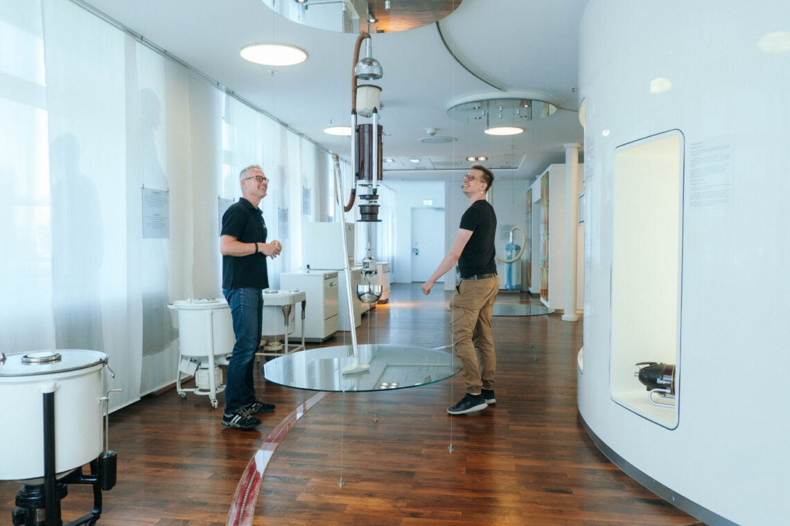 Alexx Henke und Björn Voß betrachten im Miele Museum ein historisches, von der Decke hängendes Haushaltsgerät.